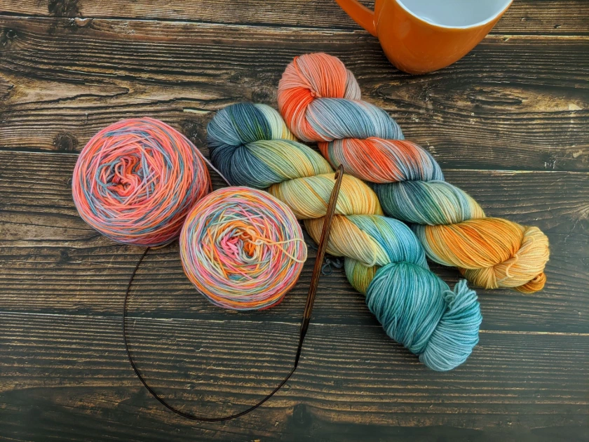 Hand-dyed yarn skeins and yarn cakes in the High Fantasy color palette, featuring bright teal, golden yellow, coral, and soft pastel tones.